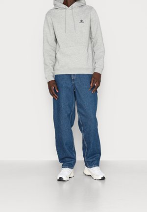 Šedá bavlněná mikina s kapucí, přední kapsou a stahovacími šňůrkami, kombinovaná s volnými modrými džínami z denimu. Nošena s bílými teniskami s černými detaily.