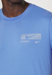 Camiseta de algodón azul con cuello redondo. Presenta un logotipo de Nike en blanco y un gráfico de texto en el área del pecho izquierdo. Textura suave.