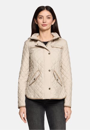 Donna che indossa una giacca trapuntata beige con bottoni a pressione e tasche con patta, abbinata a pantaloni neri, in piedi davanti a uno sfondo semplice.