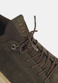 Donkerbruine sneaker met een bovenwerk van suede, een getextureerde stoffen tong en contrasterende bruine veters met metalen aglets. Accentuerende stiksels zichtbaar.