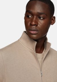 Chaqueta beige con cremallera y cuello alto, hecha de un tejido suave. Presenta un diseño sencillo con una textura sutil y detalle de cremallera visible.