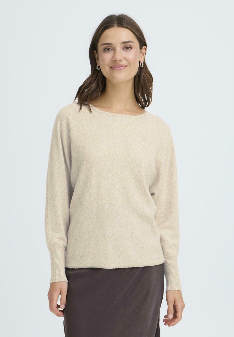 Bež pleteni pulover z okroglim izrezom, dolgimi rokavi in mehkimi teksturami, v kombinaciji s rjavim krilom, na svetli podlagi.