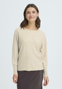 Beige stickad tröja med rundad halsringning, långa ärmar och mjuk textur, tillsammans med en brun kjol, mot en ljus bakgrund.