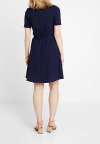 Námorně modré šaty s krátkým rukávem, který mají přiléhavý pas a rozšířenou sukni. Hladká textura a decentní švy. Nosené s béžovými sandály na bloku.