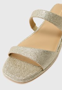 Gold glitter slide sandal featuring two wide straps, a flat sole, and a smooth beige footbed. The texture is smooth with a metallic sheen.