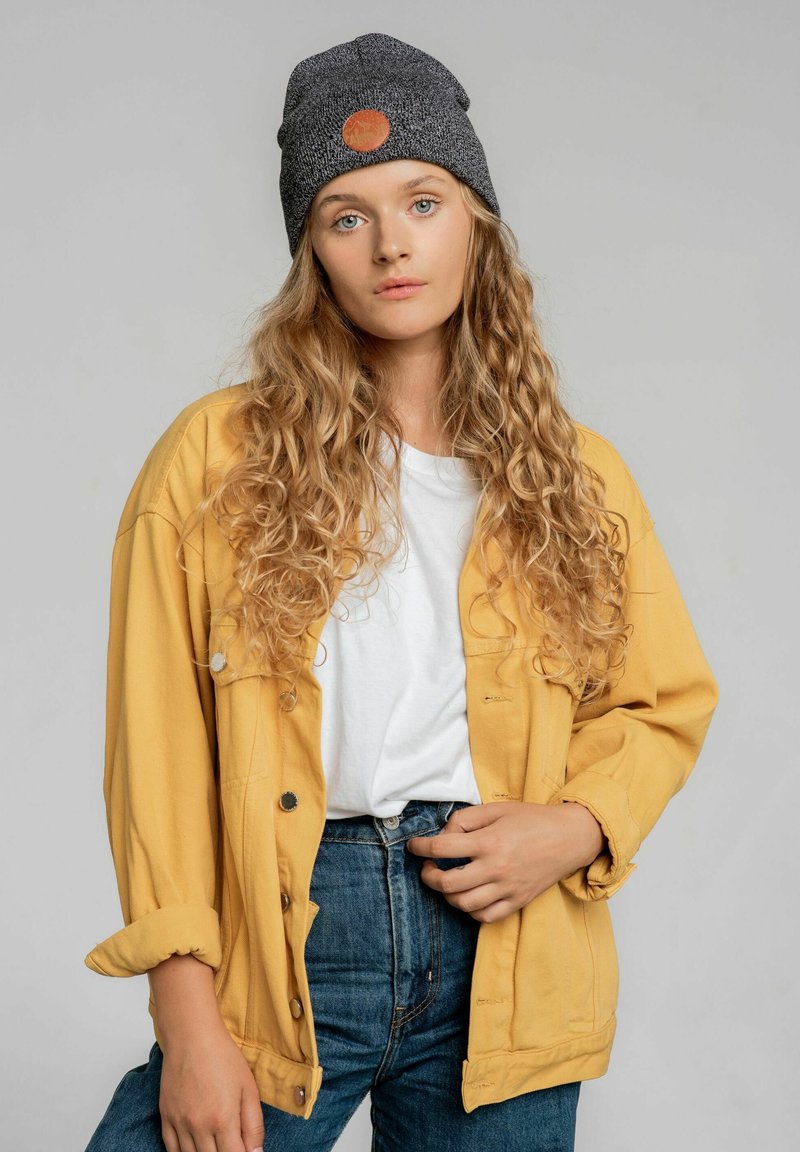 Żółta dżinsowa kurtka na białym t-shircie, zestawiona z niebieskimi dżinsami z wysokim stanem. Szary beanie z okrągłym logo dopełnia strój.