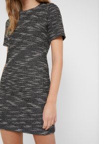 Kortärmad klänning i svartvitt texturerat tyg, med figurnära silhuett och rund halsringning, som visar ett subtilt vävt mönster.