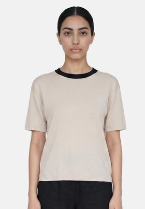 Vrouw met donker haar in een staart, gekleed in een beige shirt met korte mouwen en een zwarte kraag, staat tegen een effen lichte achtergrond.