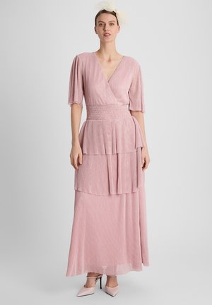 Vrouw staat en draagt een lichtroze gelaagde maxi-jurk met ellebooglange mouwen, een V-hals en bijpassende puntige hakken, tegen een effen achtergrond.