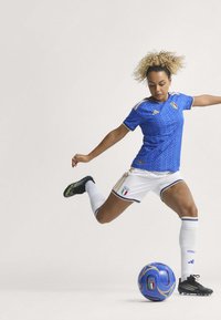 Modrý fotbalový dres s texturovaným vzorem a zlatými akcenty, doplněný bílými šortkami a černými kopačkami. V blízkosti se nachází modrý fotbal.