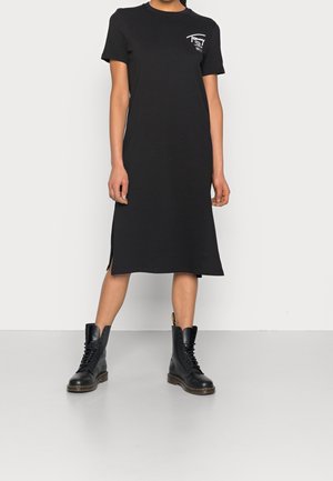 Person iført en sort midilang T-shirt-kjole med en lille hvid logo på brystet, kombineret med sorte snørestøvler.