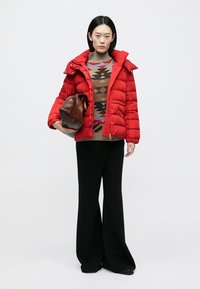 Rote Steppjacke mit Kapuze, strukturierter Oberfläche und goldenen Beschlägen, getragen über einem gemusterten Strickpullover und kombiniert mit schwarzen, weitem Hosen.