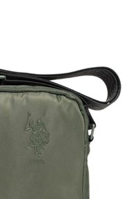 Oliwkowa torebka na ramię z wypukłym logo gracza polo USPA oraz czarnym regulowanym paskiem na białym tle.