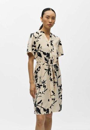 Mujer con vestido beige hasta la rodilla con estampado floral negro, mangas cortas, botones delanteros y cinturón anudado en la cintura, posando frente a un fondo liso.