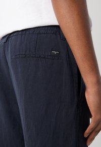 Marineblauwe broek gemaakt van lichte stof, met een achterzak en een klein merkteken. Glad oppervlak met een relaxte pasvorm.