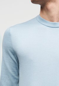 Ανοιχτό μπλε, εφαρμοστό πουλόβερ με ομαλή υφή και ριμπ στο λαιμό. Διαθέτει μακριά μανίκια και διακριτικό ράψιμο κατά μήκος της ραφής.