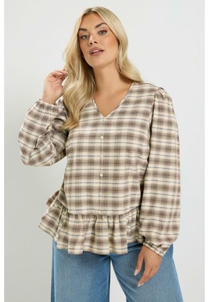 Frau mit langen blonden Haaren, die eine beige-braune karierte, geknöpfte Bluse mit Rüschen Saum und blaue Jeans vor einem einfachen Hintergrund trägt.