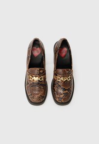 Καφέ μοκασίνια με σχέδιο από δέρμα φιδιού, διακοσμημένα με μια χρυσή αλυσίδα σε σχήμα καρδιάς και μαύρη εσωτερική επένδυση που διαθέτει σχέδιο κόκκινης καρδιάς. Πλατιά σόλα.