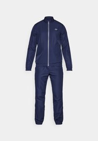 Mørkeblå træningsdress lavet af glat stof, der har en lynlåslukket jakke med ståkrave og elastiske manchetter samt matchende bukser med elastisk talje.