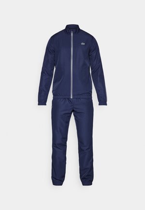 Survêtement bleu marine en tissu lisse, comprenant une veste zippée avec un col montant et des poignets élastiques, ainsi que des pantalons assortis à taille élastique.