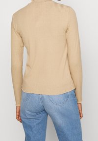 Beige gestreiftes Langarmoberteil mit einem Mock-Neck und einem leicht geschwungenen Saum, kombiniert mit hellblauen Jeans. Glatte Textur und figurnahe Passform.