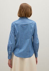 Donna con capelli castani corti, che indossa una camicia a righe blu e bianca con maniche lunghe e una gonna beige, girata di spalle su uno sfondo semplice.
