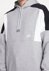 Huppari harmaa, jossa musta-valkoista väriestetiikkaa. Varustettu nyöreillä, kengurutaskulla ja pienellä valkoisella Nike-logolla rinnassa.