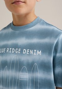 Mėlyna tie-dye marškinėliai, pagaminti iš medvilnės, su balta tekstu "BLUE RIDGE DENIM" ir banglenčių grafika, laisva forma.