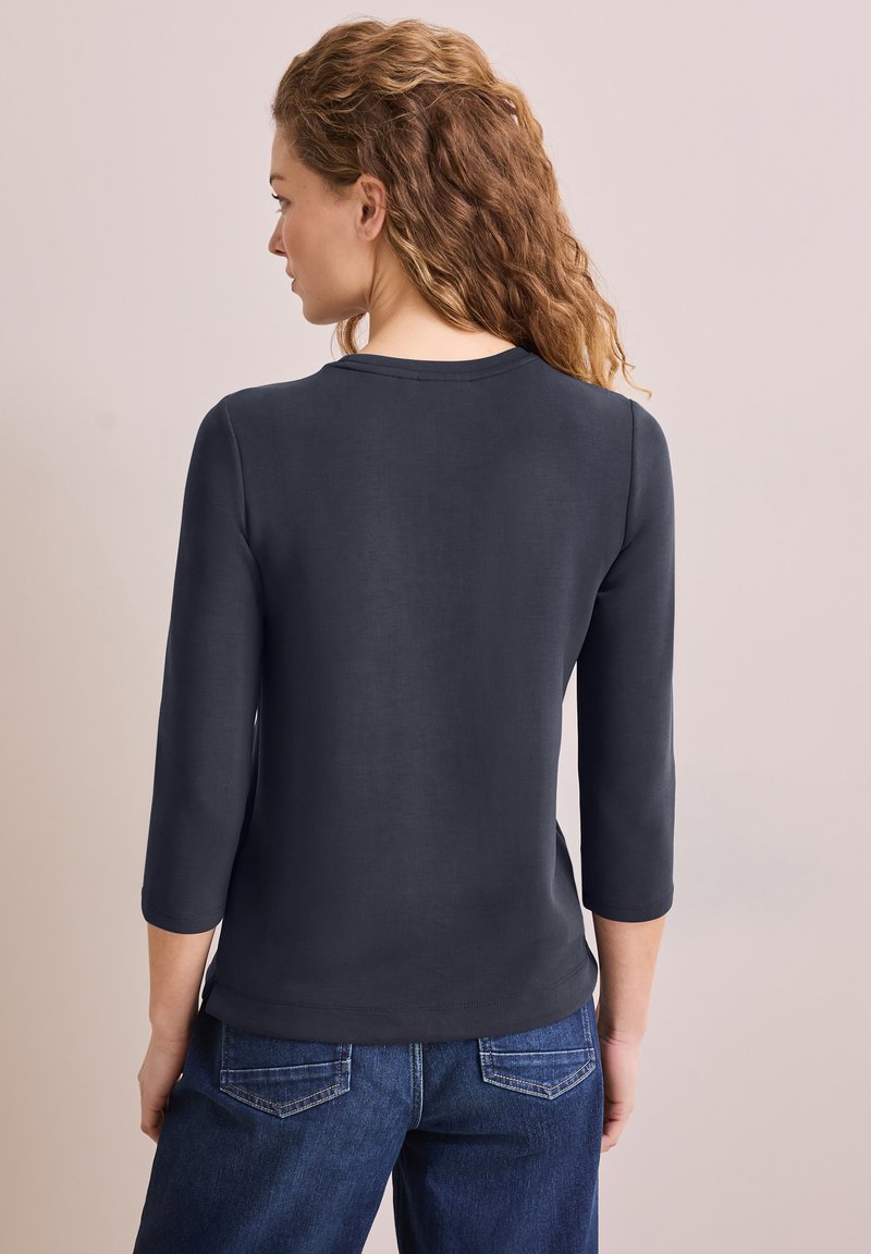 Dunkelblaues Oberteil mit drei Viertelärmeln, runder Ausschnitt, glatter Textur und leichtem Stretch. Kombiniert mit dunkelblauen Jeans.