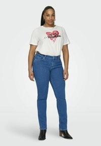 Tricou alb cu un grafic în formă de inimă roșie și text negru, asortat cu blugi skinny albaștri și bocanci negri cu toc.