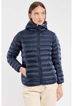 Femme portant une doudoune matelassée bleu marine avec capuche, fermeture éclair à l’avant et poches latérales, debout avec une main dans la poche.