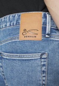 Džínové kalhoty světle modré barvy s koženou nášivkou na pase, která obsahuje logo nůžek a název "DENHAM" v debossed provedení.