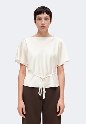 Femme aux cheveux courts et foncés portant un chemisier blanc à manches courtes avec une ceinture à la taille et un pantalon marron foncé, debout devant un fond uni.