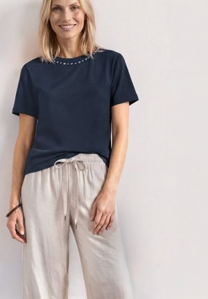 Femme souriante aux cheveux blonds porte un t-shirt bleu marine avec de petites décorations près de l'encolure et un pantalon beige clair à cordon.