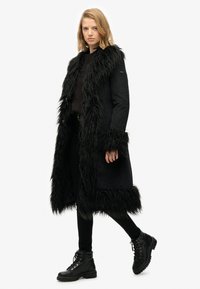 Sort lang frakke med imitation pelskant, med lige snit, åben front og strukturerede ærmer. Båret over en mørk sweater og sorte bukser.
