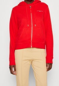 Sweat à capuche zippé rouge avec capuche, poches avant et logo brodé. Tissu doux avec une coupe courte et des poignets et ourlet côtelés.