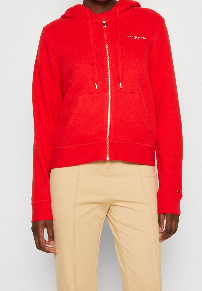 Sweat à capuche zippé rouge avec capuche, poches avant et logo brodé. Tissu doux avec une coupe courte et des poignets et ourlet côtelés.