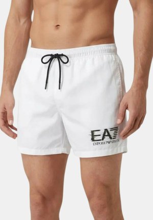 Costume da bagno bianco da uomo con coulisse nera e logo EA7 Emporio Armani sulla coscia sinistra, indossato da un modello senza maglietta.