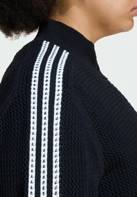 Čierny pletený sveter s vysokým golierom, ktorý má tri vertikálne biele pruhy pozdĺž ramien a štruktúrovanú tkaninu.