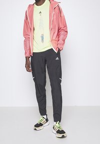 Leichte rosa Jacke, neon-grünes T-Shirt mit Grafik, schwarze Hose mit reflektierenden Streifen und schwarze Sneakers mit neonfarbenen Schnürsenkeln.