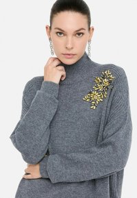Vrouw met ongelijke ogen draagt een grijze turtleneck trui met gouden en zwarte bloemversieringen en lange zilveren oorbellen, terwijl ze poseert.