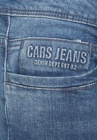 Vaqueros de mezclilla azul con una superficie texturizada, que cuentan con un bolsillo trasero cosido y una etiqueta tejida que dice "CARS JEANS" con texto adicional debajo.