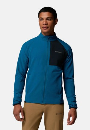 Mann trägt eine blaue Zip-up-Jacke mit schwarzer Brusttasche und beigefarbenen Hosen, steht vor einem schlichten hellen Hintergrund.