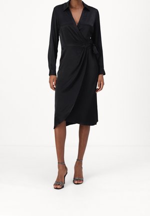 Kobieta ubrana w czarną, długorękawową sukienkę kopertową z kołnierzykiem, wiązaną w talii, z asymetrycznym dołem, w zestawieniu z czarnymi sandałami na wysokim obcasie z paskami.