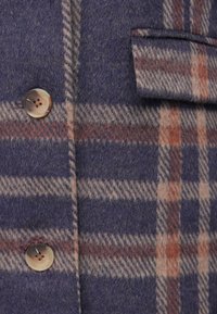 Gros plan sur un tissu en laine à carreaux violet et orange avec deux boutons ronds et un détail de poche à rabat.