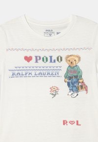 Biała koszula z kolorowym haftowanym tekstem "POLO RALPH LAUREN", pluszowy miś w casualowych ubraniach trzymający torbę oraz małe motywy kwiatowe.