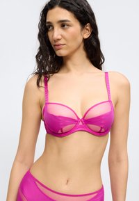 Γυναίκα με μακριά σκούρα μαλλιά που φοράει ένα φωτεινό ροζ διάφανο σουτιέν και αντίστοιχο εσώρουχο με ψηλή μέση, σε απλό γκρι φόντο.