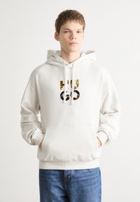 Sudadera blanca con capucha, bolsillo frontal y el logo "HUGO" en dorado y negro. Fabricada en tela suave, con un ajuste holgado y cordones.
