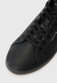 Deportiva de cuero negro con punta redonda, superficie texturizada, seis ojales y cordones planos. Diseño simple con un logotipo minimalista en el costado.