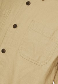 Camicia button-up beige con una singola tasca sul petto, bottoni marrone scuro, texture del tessuto liscia e design classico del colletto.